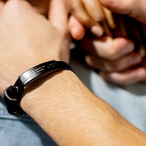Puede incluir: Un brazalete de cuero negro con una banda de metal plateado grabada con un patr&oacute;n de l&iacute;neas. El brazalete se lleva en la mu&ntilde;eca.
