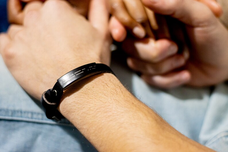 Puede incluir: Un brazalete de cuero negro con una banda de metal plateado grabada con un patr&oacute;n de l&iacute;neas. El brazalete se lleva en la mu&ntilde;eca.