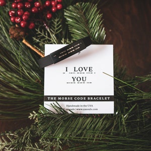 Puede incluir: Un brazalete de cuero negro con puntos dorados que deletrean "I LOVE YOU" en c&oacute;digo Morse. El brazalete est&aacute; sobre una tarjeta blanca con el texto "THE MORSE CODE BRACELET Handmade in the USA casouls.com www.casouls.com".