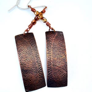 May include: A pair of copper earrings with a hammered texture. The earrings are rectangular in shape and have a silver hook fastening. The earrings are accented with a small cluster of amber-coloured beads.