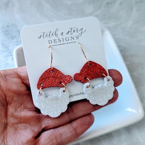 May include: Red and white glitter Santa Claus earrings with gold hooks. The earrings are on a white card with the text "stitch a story DESIGNS custom embroidery & accessories".