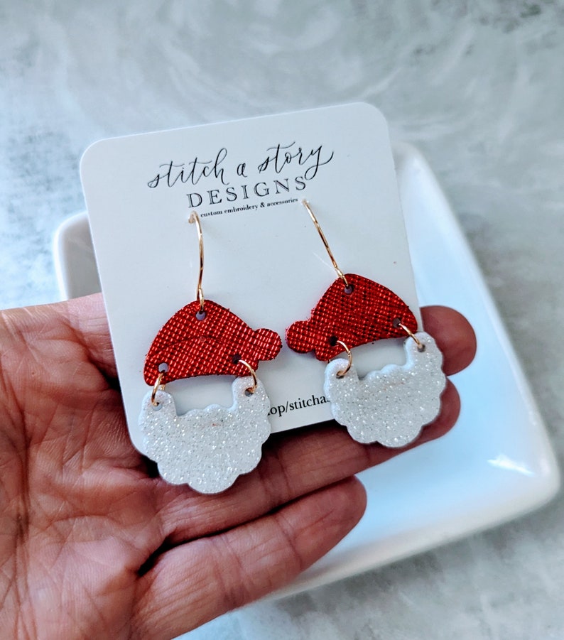May include: Red and white glitter Santa Claus earrings with gold hooks. The earrings are on a white card with the text "stitch a story DESIGNS custom embroidery & accessories".