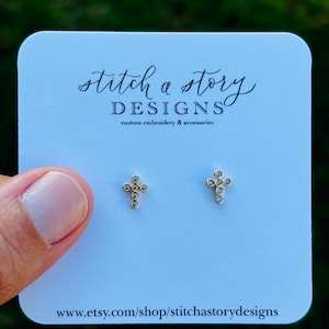 May include: A pair of gold-colored cross stud earrings with small, clear gemstones. The earrings are displayed on a light blue card with the text "stitch a story DESIGNS custom embroidery & accessories".
