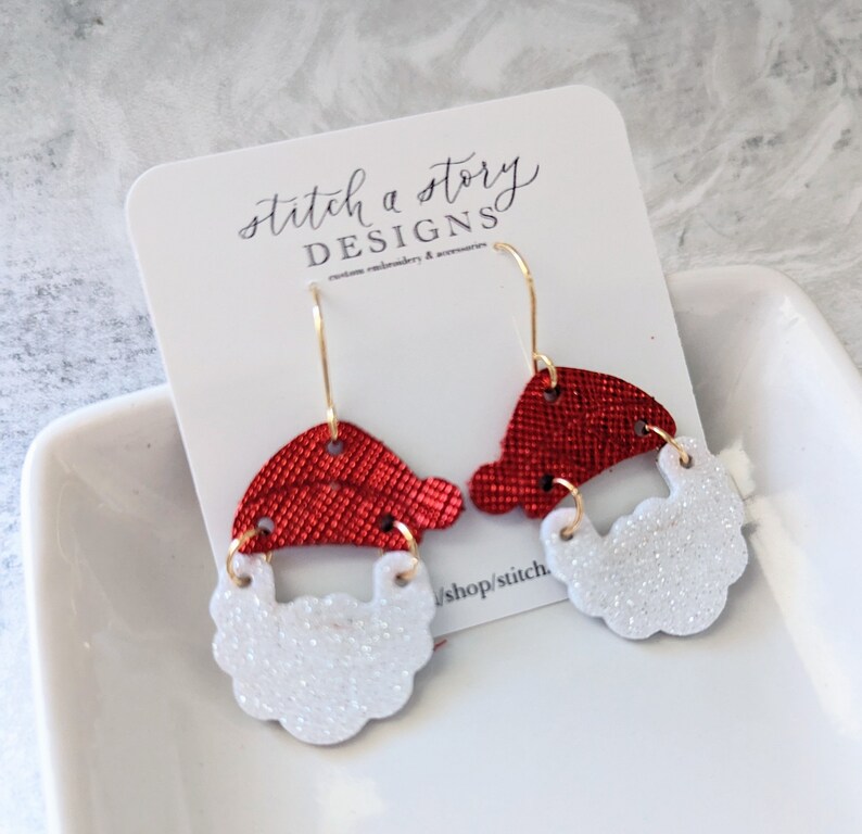 May include: A pair of red and white Santa Claus earrings. The earrings are made of glitter and have a gold hook.