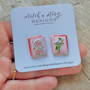 May include: Two enamel pins on a white card. One pin features a pink background with two floral designs and the number 2. The other pin has a pink background with a bird on a branch and the number 1. The card reads "stitch a story DESIGNS".