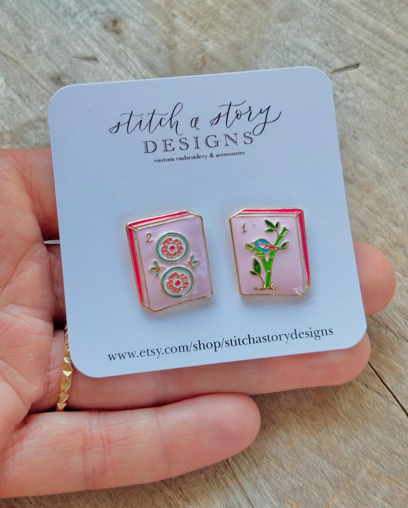 May include: Two enamel pins on a white card. One pin features a pink background with two floral designs and the number 2. The other pin has a pink background with a bird on a branch and the number 1. The card reads "stitch a story DESIGNS".