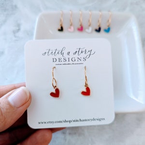 May include: Red heart dangle earrings with gold hooks. The earrings are on a white card with the text "stitch a story DESIGNS custom embroidery & accessories www.etsy.com/shop/stitchastorydesigns"