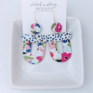 Puede incluir: Un par de pendientes abstractos coloridos con aros dorados. Los pendientes presentan una mezcla de patrones rosas, azules, verdes y blancos. La parte superior de los pendientes tiene un diseño de lunares. Una tarjeta blanca con el texto "stitch a story DESIGNS" está detrás de los pendientes.