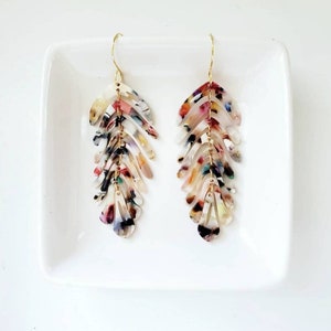 May include: A pair of long, colorful, multi-layered, dangle earrings with a gold hook. The earrings are made of a translucent material with a marbled pattern in shades of white, pink, green, and blue.