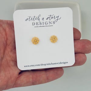 May include: A pair of gold-colored stud earrings with a clover-like design. The earrings are displayed on a white card with the text "stitch a story DESIGNS custom embroidery & accessories" and a website address.