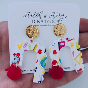 May include: A pair of white acrylic earrings with a school-themed pattern. The earrings are shaped like an arch and have a red apple charm hanging from the bottom. The earrings are accented with gold-toned metal.