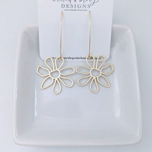 May include: A pair of gold-colored flower-shaped earrings with long, thin hooks. The earrings are displayed on a white card with the text "stitch a story DESIGNS" and a white ceramic dish.