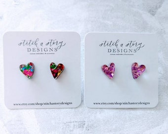 May include: Two pairs of heart-shaped earrings. The first pair is pink with glitter and the second pair is multi-colored with a floral pattern. Both pairs are on white cards with the text "stitch a story DESIGNS" and a website address.
