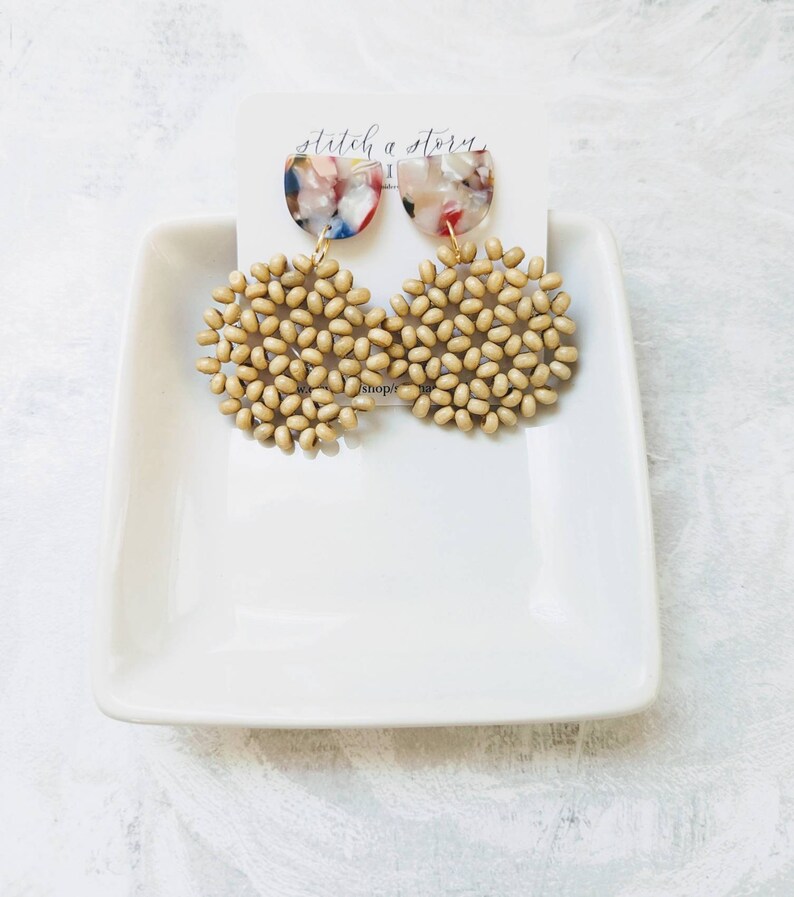 Peut inclure: Une paire de boucles d'oreilles avec un groupe de perles rondes en bois beige et un pendentif en r&eacute;sine translucide multicolore en demi-cercle. Les boucles d'oreilles sont sur une plaque carr&eacute;e blanche avec le texte "stitch a story" sur une carte blanche derri&egrave;re elles.