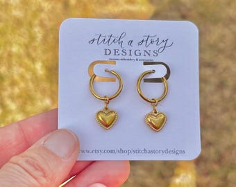 Puede incluir: Un par de pendientes de aro dorados con colgantes en forma de corazón. Los pendientes se muestran en una tarjeta blanca con el texto "stitch a story designs custom embroidery & accessories" y una dirección web.