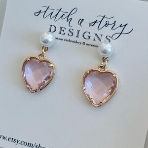 May include: A pair of pink heart-shaped earrings with gold accents and pearl accents. The earrings are on a white background with the text "stitch a story DESIGNS custom embroidery & accessories" and the website address "www.etsy.com/shop/stitchastoryd".
