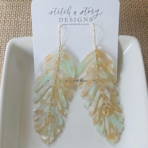 May include: A pair of leaf-shaped earrings with a gold-colored chain and ear wire. The earrings are made of a translucent material with a mix of light green, beige, and white colors. The earrings are displayed on a white card with the text "Stitch a Story Designs".