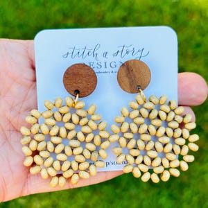 Peut inclure: Une paire de boucles d'oreilles en perles de bois avec un cercle en bois brun en haut. Les boucles d'oreilles ont la forme d'une fleur avec un motif circulaire. Les boucles d'oreilles sont sur une carte blanche avec le texte "stitch a story design embroidery & art".