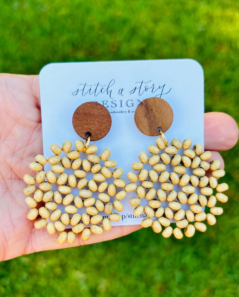 Peut inclure: Une paire de boucles d'oreilles en perles de bois avec un cercle en bois brun en haut. Les boucles d'oreilles ont la forme d'une fleur avec un motif circulaire. Les boucles d'oreilles sont sur une carte blanche avec le texte "stitch a story design embroidery & art".
