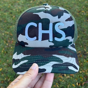 May include: Camouflage print baseball cap with the embroidered letters "CHS" in light blue. The cap features a mesh back and a curved brim. The hat is a mix of green, brown, and gray colors.
