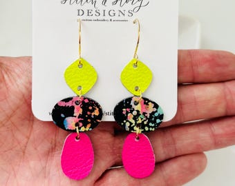May include: A pair of dangle earrings with three layers of leather. The top layer is a bright yellow square, the middle layer is a black and white splatter pattern, and the bottom layer is a bright pink oval. The earrings are hanging from gold hoops.