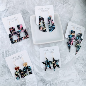 May include: Four pairs of dangle earrings with black and white paint splatter designs. The earrings are made of leather and have gold hooks. The designs include a lightning bolt, a star, an arch, and a geometric shape.