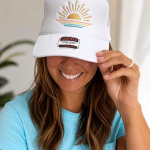 Puede incluir: Una gorra de camionero blanca con un gráfico bordado de un sol con rayos dorados, una línea de horizonte rosa y olas azules. El panel frontal muestra una pegatina rectangular con el texto OTTO WORLD FAMOUS FOAM FRONT TRUCKER.