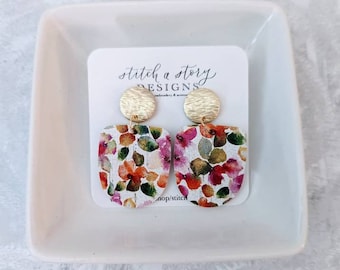 Può includere: Un paio di orecchini con stampa floreale e dettagli dorati. Gli orecchini sono su una carta bianca con il testo "stitch a story DESIGNS embroidery & more shop/stitch".