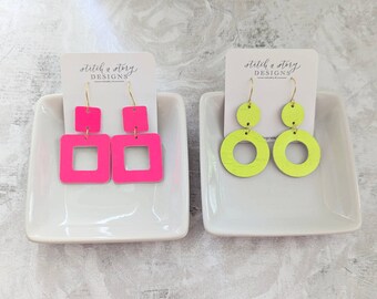 May include: Two pairs of earrings on white display stands. The earrings are made of leather and have a geometric design. One pair is neon pink and the other is neon green.
