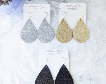 May include: Three pairs of teardrop-shaped dangle earrings with a glitter finish. The earrings are silver, gold, and black. The earrings are displayed on white cards with the text "stitch a story DESIGNS".