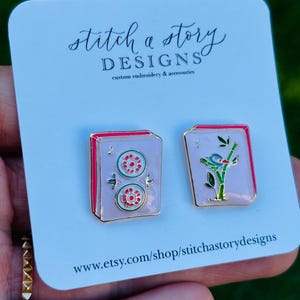 May include: A pair of enamel earrings on a white card. One earring features two red and green circles, the other a bird on a branch. The card reads "stitch a story DESIGNS custom embroidery & accessories" and the Etsy shop URL.