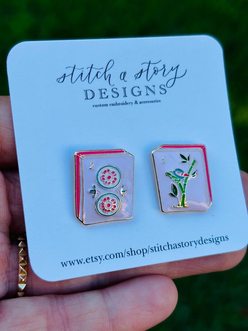 May include: A pair of enamel earrings on a white card. One earring features two red and green circles, the other a bird on a branch. The card reads "stitch a story DESIGNS custom embroidery & accessories" and the Etsy shop URL.