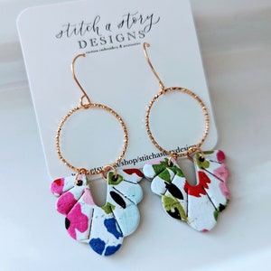 Puede incluir: Aretes de aro dorados con un diseño floral. El diseño floral está hecho de tela blanca con flores rosas, azules, verdes y rojas. Los aretes son de Stitch a Story Designs.