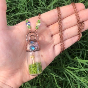 Puede incluir: Un collar de cobre con un colgante de vial de vidrio que contiene un pequeño hongo y musgo verde. El vial está coronado con una tapa de cobre con una piedra preciosa azul.