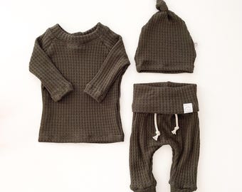 Peut inclure: Un ensemble de vêtements en maille gaufrée vert olive foncé composé d'un haut à manches longues, d'un pantalon avec une taille repliée et un cordon blanc, et d'un bonnet à nœud. Le tissu présente un motif quadrillé texturé sur l'ensemble.