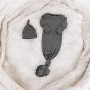 Puede incluir: Conjunto de pijama para beb&eacute; gris oscuro y gorro a juego. El pijama tiene mangas largas y un bajo anudado. Ambos art&iacute;culos est&aacute;n hechos de tejido acanalado y se muestran sobre una manta blanca y suave.