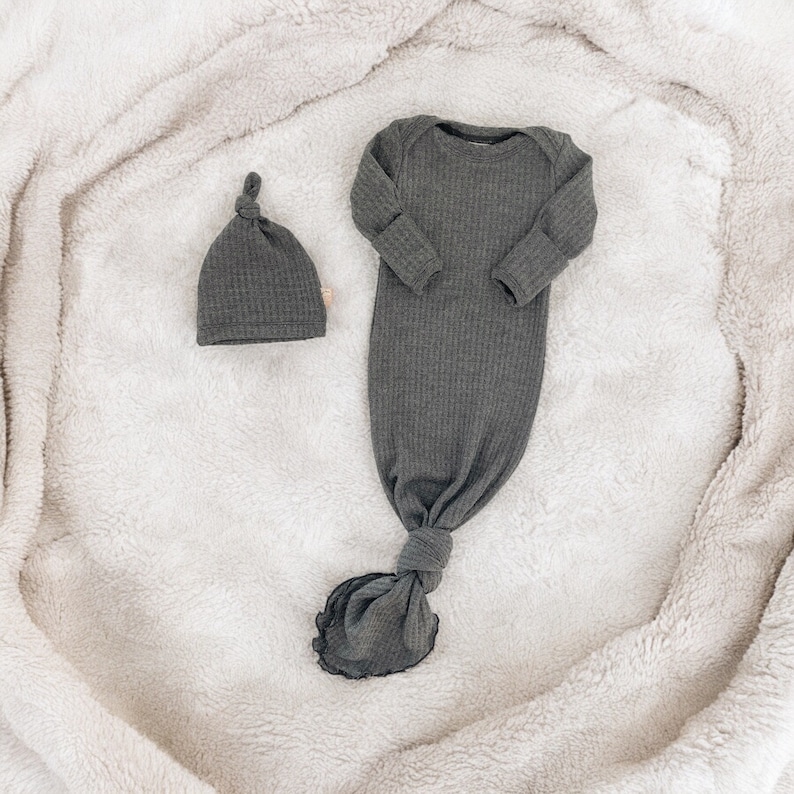 Puede incluir: Conjunto de pijama para beb&eacute; gris oscuro y gorro a juego. El pijama tiene mangas largas y un bajo anudado. Ambos art&iacute;culos est&aacute;n hechos de tejido acanalado y se muestran sobre una manta blanca y suave.