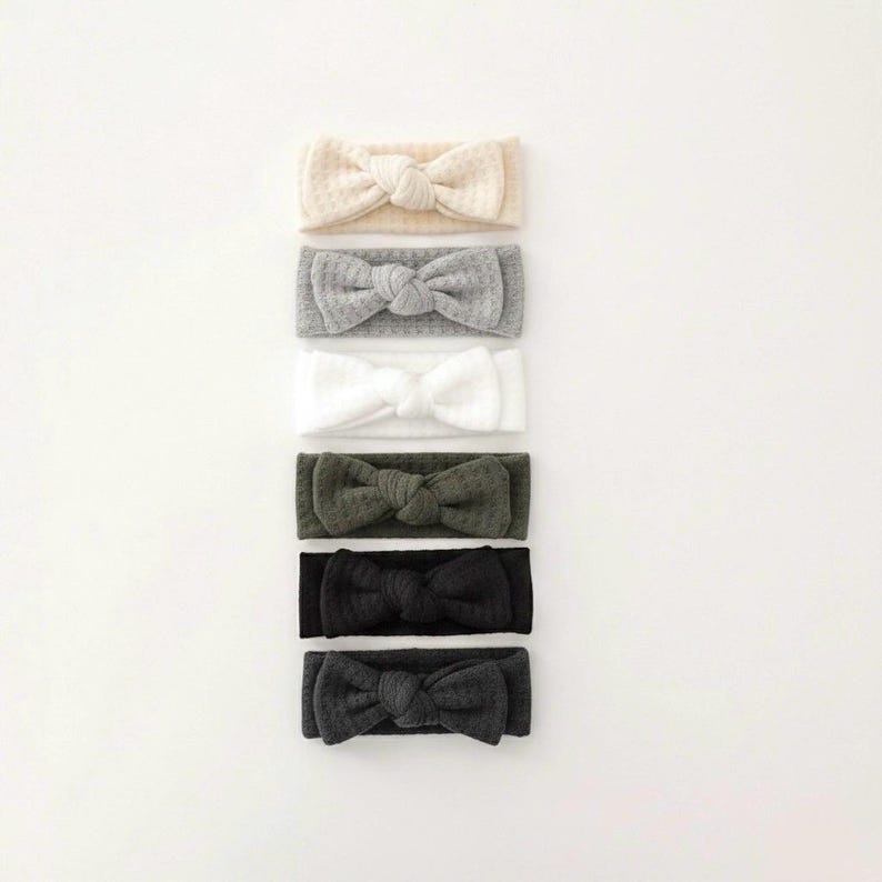 Puede incluir: Seis diademas con lazos en una variedad de colores neutros: crema, gris, blanco, verde oliva, negro y gris carb&oacute;n. Las diademas est&aacute;n hechas de una tela texturizada y est&aacute;n dispuestas verticalmente sobre un fondo blanco.