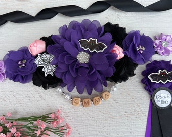 May include: A purple and black Halloween-themed baby shower sash with floral accents, a bat, and a spiderweb. The sash includes the name "EMMA" in wooden letters and a "Daddy to be" pin.