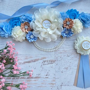 K&ouml;nnte beinhalten: Ein blaues und wei&szlig;es Band mit Blumen und einer Perlenkette, mit einer wei&szlig;en und goldenen "Mommy To Be"-Nadel und einer wei&szlig;en und goldenen "Daddy To Be"-Nadel mit einem blauen Band.