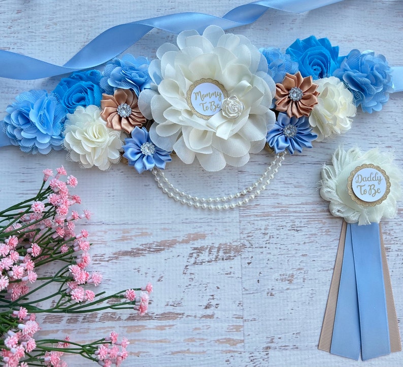 K&ouml;nnte beinhalten: Ein blaues und wei&szlig;es Band mit Blumen und einer Perlenkette, mit einer wei&szlig;en und goldenen "Mommy To Be"-Nadel und einer wei&szlig;en und goldenen "Daddy To Be"-Nadel mit einem blauen Band.