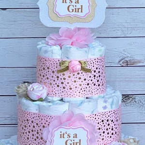 Puede incluir: Una tarta de pa&ntilde;ales de tres niveles decorada con detalles rosas y dorados. La tarta presenta cintas rosas con lunares dorados, flores artificiales y letreros que dicen "it's a Girl". Un lazo decorativo y una rosa adornan el nivel intermedio.
