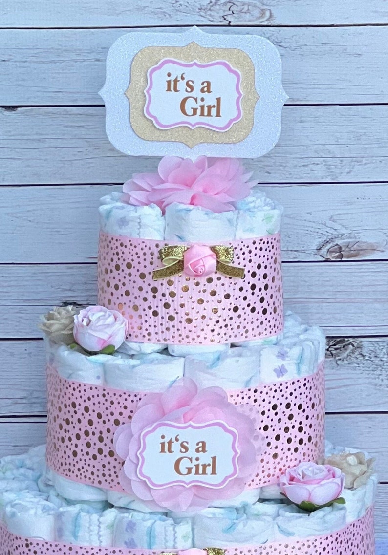 Puede incluir: Una tarta de pa&ntilde;ales de tres niveles decorada con detalles rosas y dorados. La tarta presenta cintas rosas con lunares dorados, flores artificiales y letreros que dicen "it's a Girl". Un lazo decorativo y una rosa adornan el nivel intermedio.