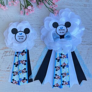May include: Two white satin flower corsages with blue and black ribbon. The corsages have a Mickey Mouse head design with a blue baby footprint in the center. One corsage says "Mommy to Be" and the other says "Daddy to Be".