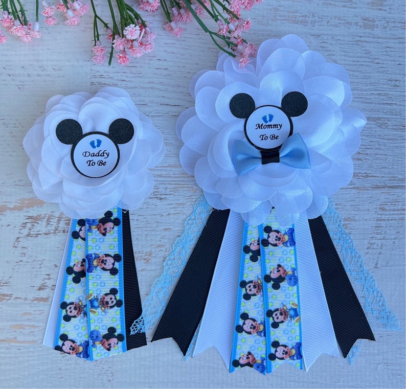 May include: Two white satin flower corsages with blue and black ribbon. The corsages have a Mickey Mouse head design with a blue baby footprint in the center. One corsage says "Mommy to Be" and the other says "Daddy to Be".