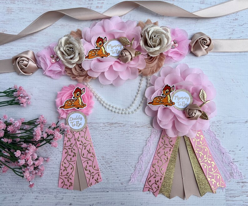 May include: Pink and gold mommy-to-be and daddy-to-be sashes with a floral design and a cartoon Bambi. The sashes have the text "Mommy to Be" and "Daddy to Be" printed on them.