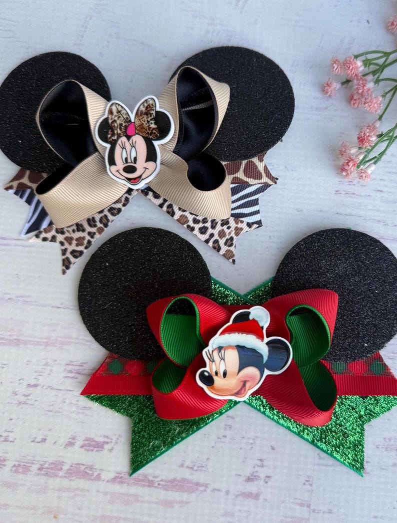 May include: Two black glitter mouse ears with bows. The first bow is brown, black, and white with a Minnie Mouse face in the centre. The second bow is red and green with a Minnie Mouse face wearing a Santa hat in the centre.