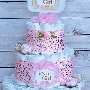 Puede incluir: Una tarta de pa&ntilde;ales de tres niveles decorada con detalles en rosa y adornos florales. La tarta presenta una banda rosa perforada alrededor de cada nivel y est&aacute; coronada con un letrero que dice "it's a Girl".