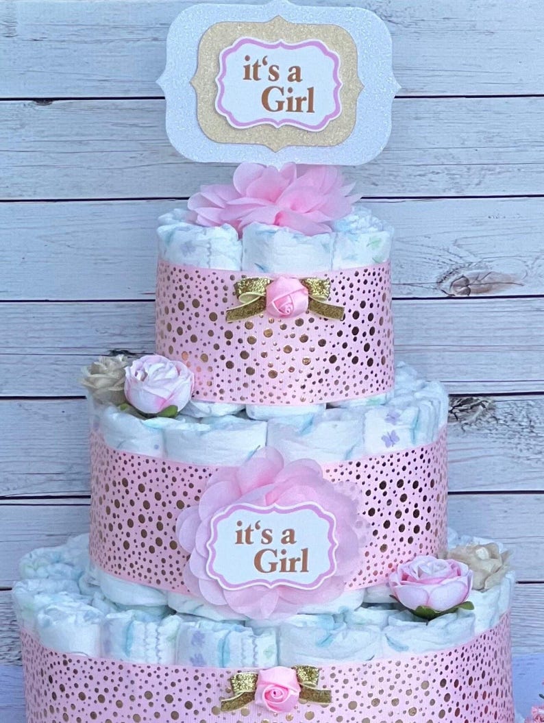 Puede incluir: Una tarta de pa&ntilde;ales de tres niveles decorada con detalles en rosa y adornos florales. La tarta presenta una banda rosa perforada alrededor de cada nivel y est&aacute; coronada con un letrero que dice "it's a Girl".