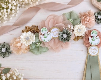 May include: A maternity sash and matching badge set featuring sage green, cream, and dusty rose fabric flowers with rhinestone centers. The sash includes a dinosaur graphic and a circular tag reading Mommy To Be, while the badge reads Daddy To Be.